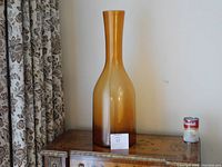 Tall amber-colored Scandinavian glass floor vase on wooden table with floral curtain background and Campbell's soup can for scale.