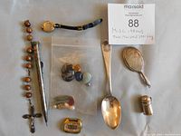 Full view of all items including the silver miniature hand mirror, rosary with wooden beads, a metal pen, small gold-colored watch, miniature metal container with lid, a silver spoon, and a plastic bag containing various beads and stones.