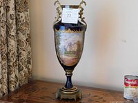 Full view of the tall porcelain vase showing its general shape, gold ornate handles and base, and the blue body with painted scenes.