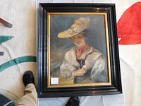 Framed oil painting featuring a woman in period clothing and a hat, holding a flower, with framing on a carpet showing part of a foot for scale