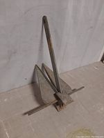 Metal boat anchor placed upright against a wall, front perspective showing the two triangular flukes and long shank with attachment hole.