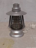 Side angled view of silver Sunbeam bug zapper showing protective cage, controls on base, power adapter plug on surface.