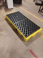Drum spill deck showing top view with black grate surface on yellow base on warehouse floor.