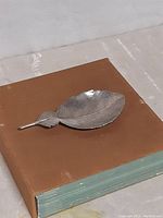 Feather-shaped pewter serving tray shown on top of a closed brown book, highlighting size and detail.