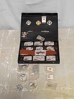 Photo of open case showing multiple badges on upper section, several engraved belt buckles with leather belt below, and loose pins positioned in front of case.