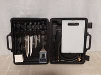 Top view of black plastic carrying case open to show 4 knives, tongs, spatula, grill brush, cutting board, and hose