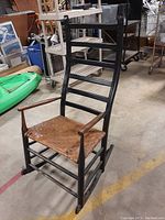 Side angled view of black wooden rocking chair with woven seat and natural wood armrests