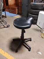 Black adjustable rolling stool with saddle-shaped faux leather seat, five-legged plastic caster base, photographed from front angle on concrete floor.