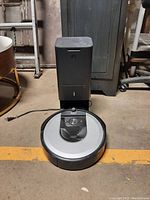 Front view of iRobot Roomba vacuum cleaner docked in charging and self-emptying base station, showing circular vacuum with control panel and base unit on floor.