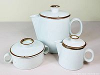 Photo showing three pieces: teapot, creamer, and sugar bowl with lids, all with brown trim and oval handles on white stoneware.