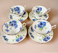 Four white tea cups with blue cornflower floral patterns and matching saucers arranged on a flat surface, showing delicate gold trim on rims and handles.