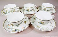 Five white porcelain tea cups and matching saucers arranged side by side with detailed colorful bird and flower patterns and gold rim trim.