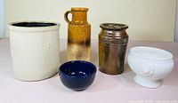 Photo showing all five pottery and stoneware pieces arranged on a white surface: white jar, brown handled vase, brown jar, blue bowl, and white footed dish.