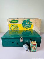 Front view of green Coleman camp stove model 421A with original worn box behind it, along with the Coleman filter funnel and its box in front.