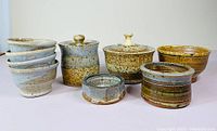 Photo showing full lot of five light blue bowls, two lidded bowls (one blue-gray, one speckled beige), one medium yellow-brown bowl, and one small banded bowl.