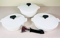 Three white Corning Ware buffet server dishes with lids and black interchangeable handle laid out on a flat surface showing the lot contents.