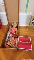 American Girl Isabelle doll lying in an open cardboard box with two books and a red and white box beside it.