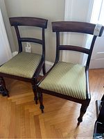 Two wood dining chairs with green striped upholstery seats showing front and side angles on wood floor.