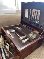 Open wooden canteen displaying multiple pieces of ornate silver flatware organized in fabric-lined slots.