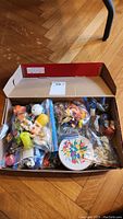 Box full of assorted keychains, toys, pins, and knick knacks packed mostly in plastic bags on a wooden floor.
