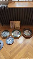 Six collector's plates and two framed pieces arranged on the floor, some leaning against radiator, showing diversity in plates and framed floral arts