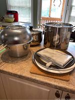 Image showing the full assortment on the kitchen counter including pots, strainers, cheese board with knife, serving plates, and wooden cutting board.