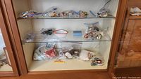 Photo shows a wooden display cabinet with three shelves containing clear plastic bags filled with assorted dolls, doll clothes, buttons, and patches.
