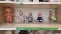 Five vintage plastic baby dolls arranged on a shelf, includes one Kewpie doll and four others with clothing, showing overall set condition and style.