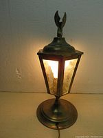 Front view of the antique brass hexagonal table lamp switched on, showing illuminated warm light through textured glass panels with eagle finial on top.