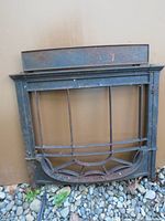 Cast iron woodstove window with glass, rectangular frame with metal bars, decorative curved base, resting on gravel with brown backdrop.