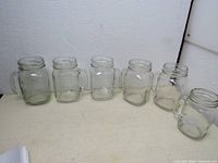 Six clear glass drinking jars with handles in a line, five same size and one smaller.