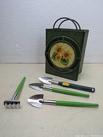 Photo presenting the metal basket with sunflower design alongside four small gardening tools with green and dark green plastic handles and metal heads.