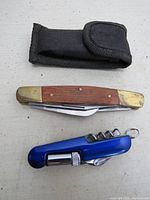 Two pocket tools side by side on light surface with black fabric carrying case above: brass-ended folding pocket knife with wooden inlays; blue multitool with shiny blue metal casing.