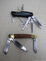 Two pocket tools laid flat showing blades extended: a brass-ended wooden folding knife with three blades and a black plastic multitool with 11 tools marked 'Year 2000'.