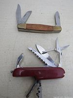 Two pocket knives displayed, one with brass ends and wooden handle and three blades, the other a red multitool multitool with multiple tools extended.