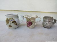 Front view of three pottery pieces: FULTON marked pitcher, CABERNET hand painted grape pitcher, and brown speckled mug