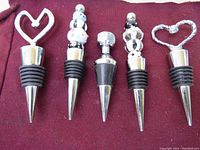 Five metal bottle stoppers with rubber rings and decorative tops including twisted heart, flat heart, nut shape, and bead decorations on a burgundy cloth background.