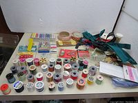 Overview photo of the sewing box material showing numerous spools of thread in various colors, zippers, buttons, tapes, and a small lock on a white table surface.