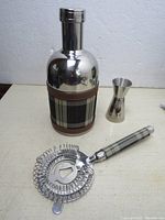 Photo shows stainless steel cocktail shaker with plaid fabric wrap, double jigger, and cocktail strainer.