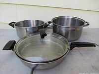 Photo showing three heavy stainless steel cookware items: one frying pan with glass lid and two smaller pots without lids arranged on grey surface