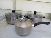 Three stainless steel pots with lids: two large with dual side handles and one smaller with a long black handle. Pots are displayed on an outdoor surface.
