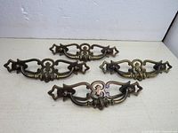Four ornate vintage brass drawer pullers arranged evenly on a flat surface, showing front and angled view for detail.