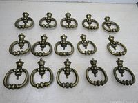 Fifteen vintage brass drawer/cabinet pullers arranged in rows on a white surface showing the front detail of each puller.