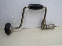 Side view of vintage hand drill showing curved metal body, black wood handle and knob, metal chuck.