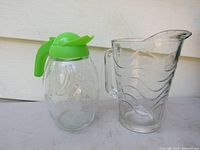 Photo showing both glass jugs side by side. Left jug has green plastic lid and handle; right jug is thick glass with wave pattern and spout.
