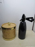 Photo showing vintage Kraftware USA gold-tone metal ice bucket with lid and dark blue glass soda bottle with dispenser next to each other on a flat surface against a white wall.
