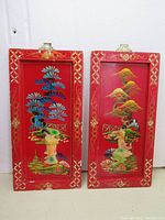 Two red wood panels displayed together showing hand painted colorful landscapes and raised 3D woman figures on each.