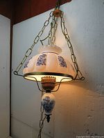 Vintage milk white glass hurricane hanging light shown lit against a wall, showing blue floral decorations and metal chain mount.