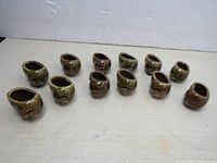Photo showing all 12 ceramic escargot snail shaped cups arranged in two rows on a white surface, displaying the size and shape of each cup with a glaze in various earthy tones.