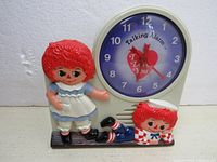 Front view showing Raggedy Ann and Andy figures flanking the circular clock face with heart-shaped graphic and 'Talking Alarm' text on purple background.